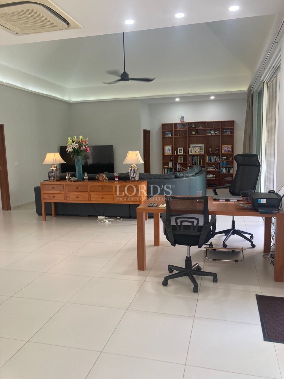 Spacious open-plan living room with home office setup, modern lighting, ceiling fan, and built-in shelving.