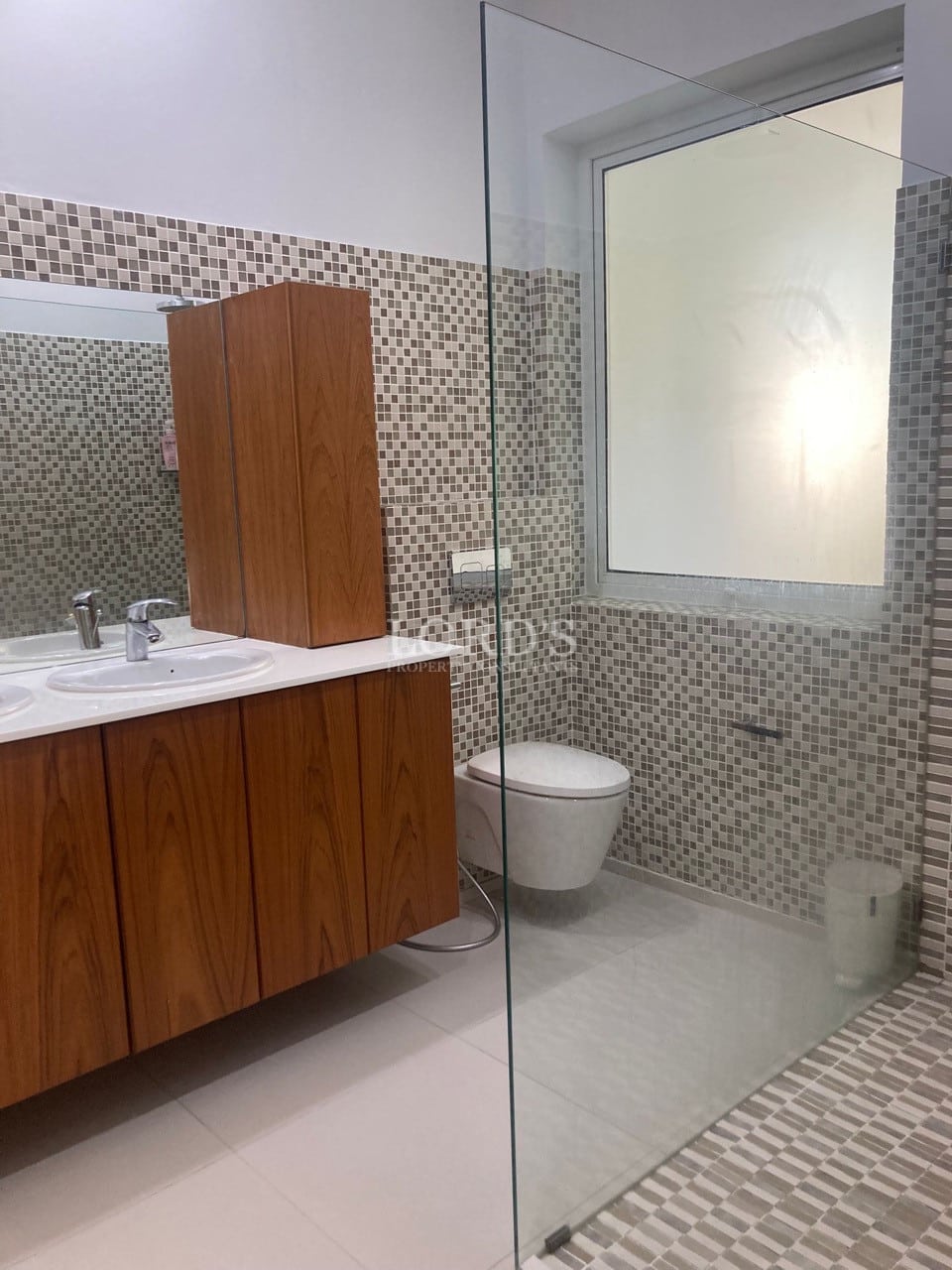 Modern bathroom with wooden vanity, mosaic tile walls, wall-hung toilet, and glass shower enclosure.