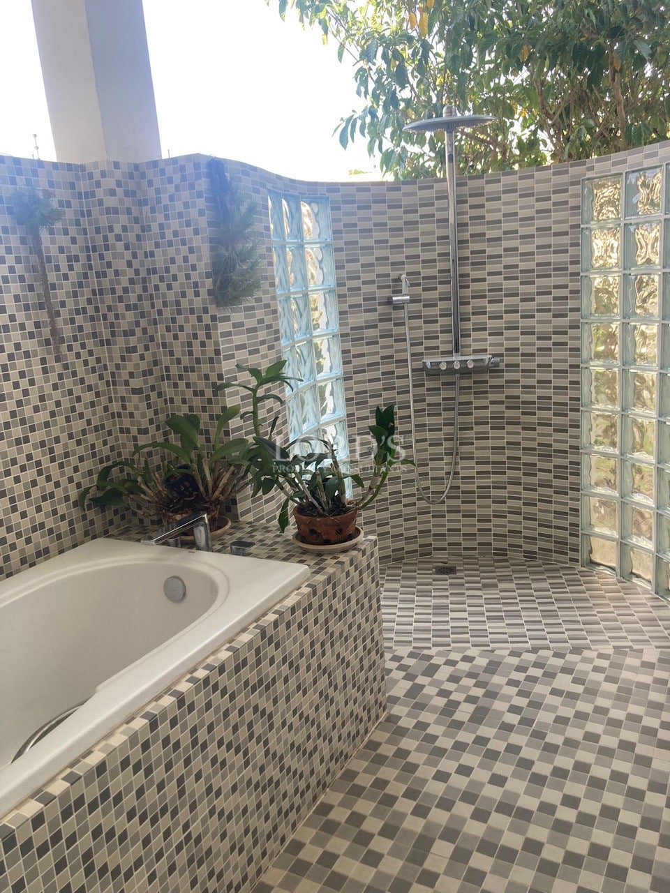 Outdoor bathroom with mosaic-tiled bathtub, walk-in rain shower, and glass block walls surrounded by greenery.