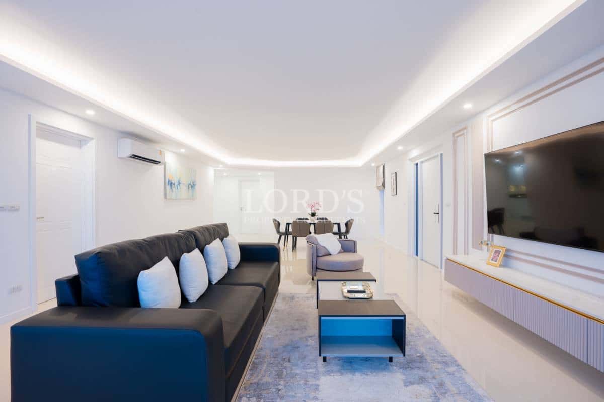 Long open-plan living and dining area with modern sofa, feature TV wall, recessed ceiling lighting, and minimalist décor.