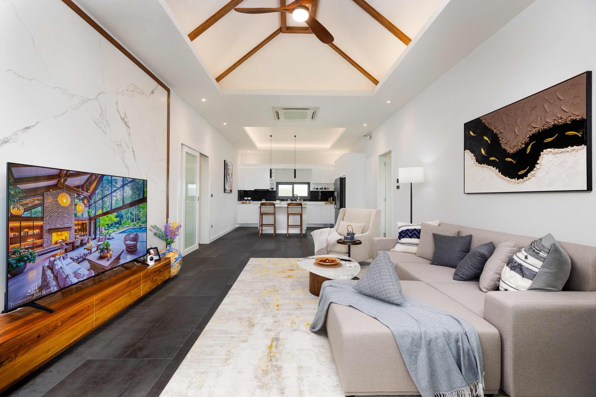 Modern open-plan living room with high ceiling, wooden roof details, comfortable sofa, and kitchen in the background.