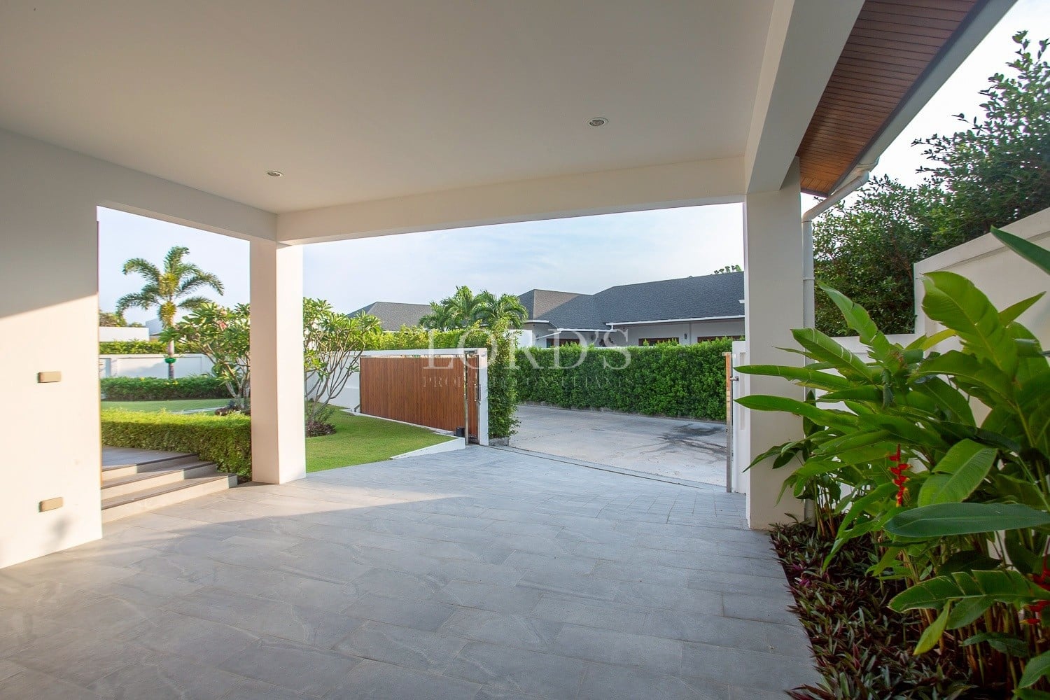 Covered driveway of a modern luxury villa with open gate, tropical landscaping, and spacious parking area.
