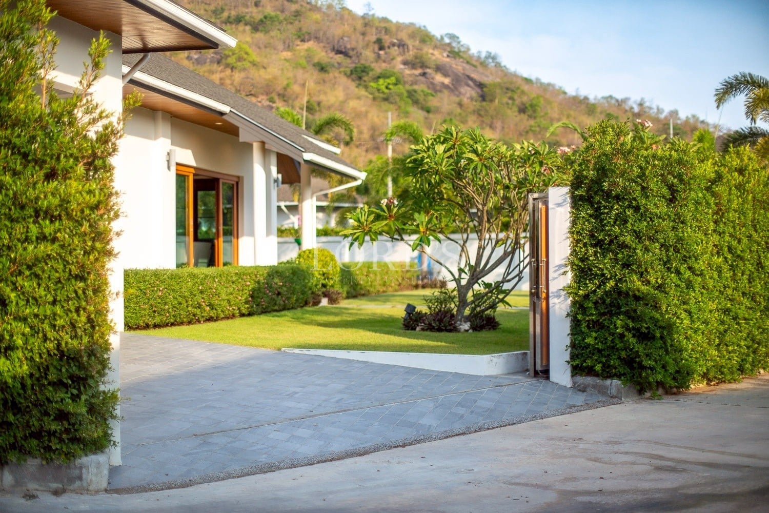 Modern luxury villa entrance with open gate, landscaped garden, and paved driveway in a tropical residential setting.