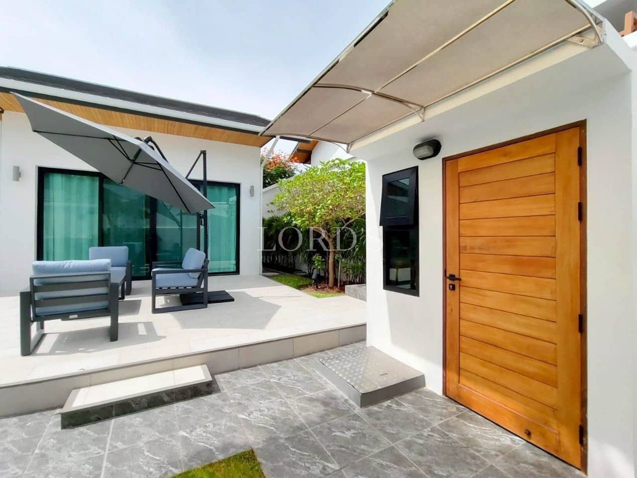 Modern villa entrance with a wooden door, shaded patio seating, and sliding glass doors.