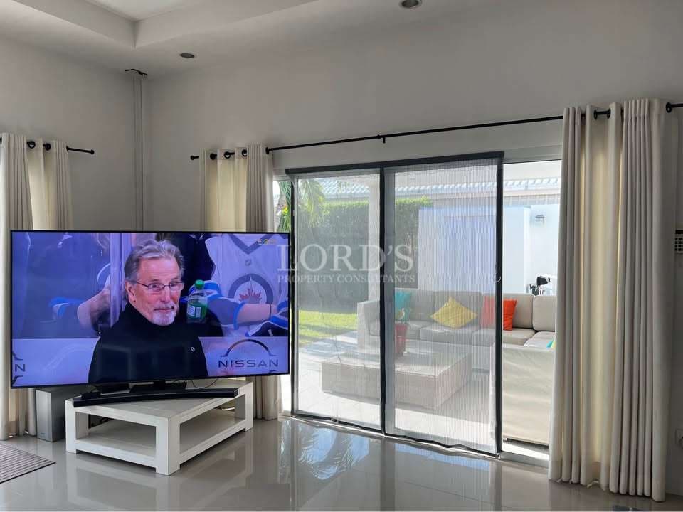 Modern living room with large flat-screen TV, sliding glass doors, sheer curtains, glossy tiled floor, and outdoor patio view