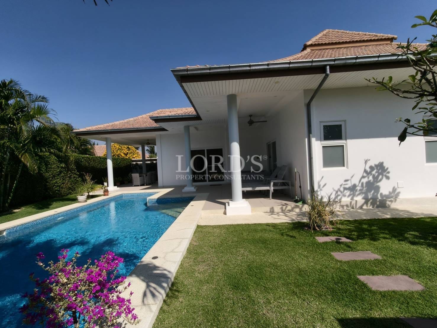 Luxury villa exterior with private swimming pool, covered patio, white façade, and manicured garden under clear blue sky.