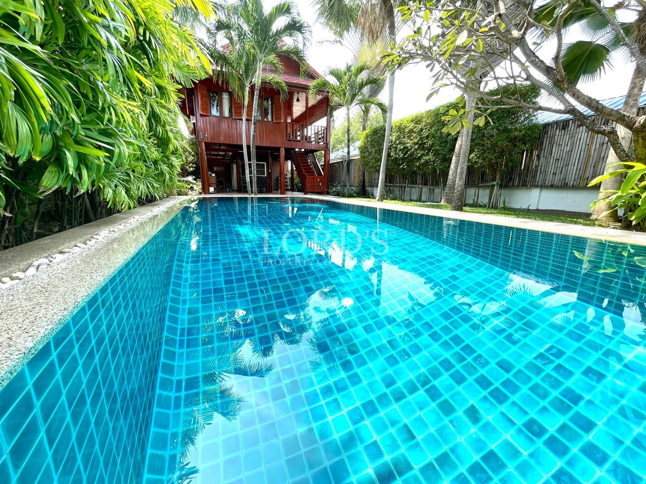 Private swimming pool with blue mosaic tiles facing a two-story wooden villa surrounded by palm trees.