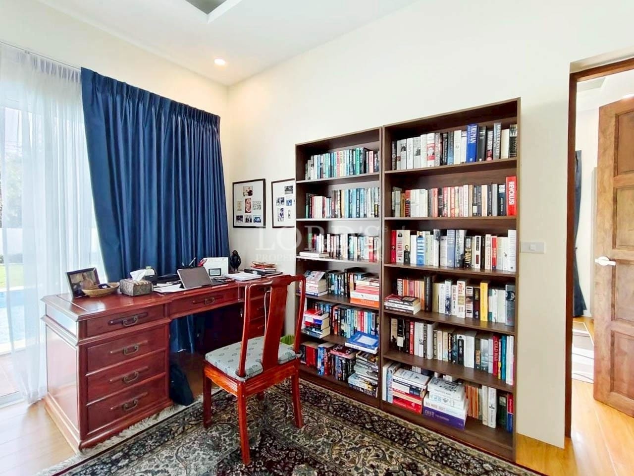 Home office with wooden study desk, chair, and full-height bookshelf filled with books near a window with blue curtains.