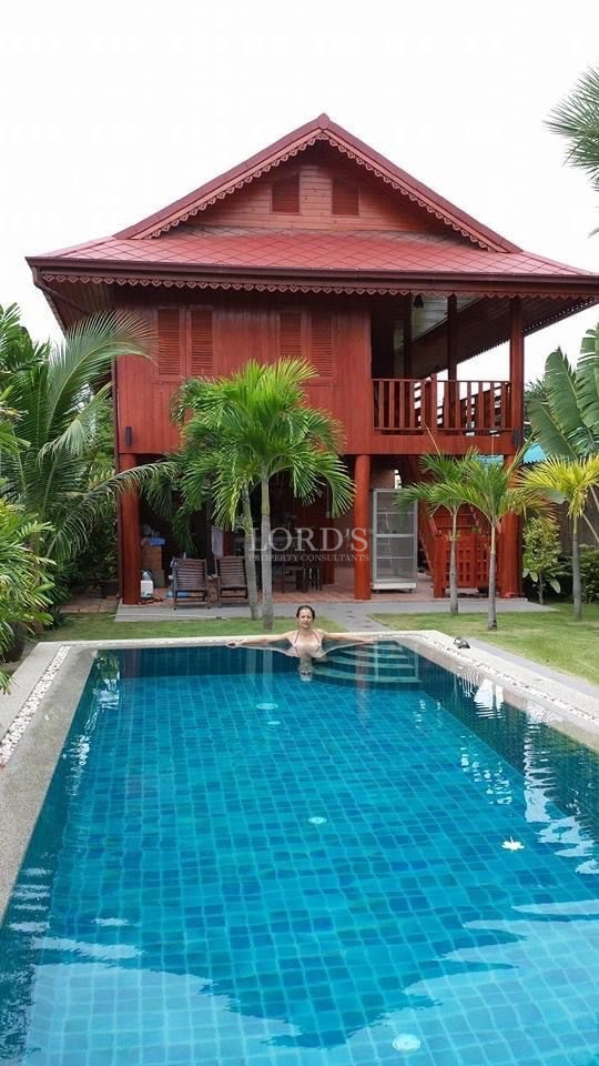 Swimming pool in front of a two-story wooden villa surrounded by palm trees, with a person relaxing in the pool.
