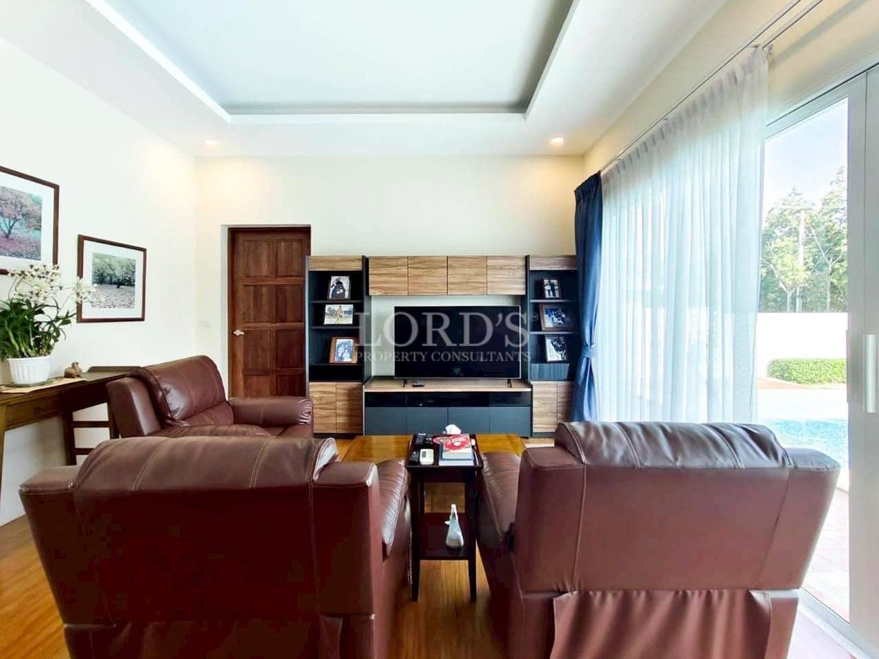Modern living room with leather recliner sofas, wooden TV unit, bookshelf storage, and pool view through large glass windows.