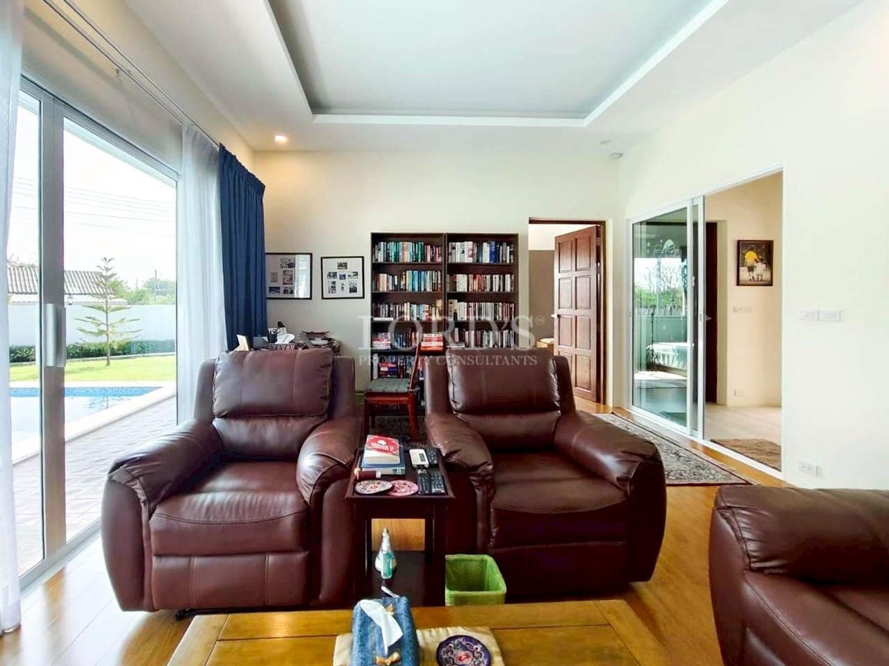 Elegant living room with leather recliner chairs, wooden flooring, bookshelf, and poolside view through glass doors.