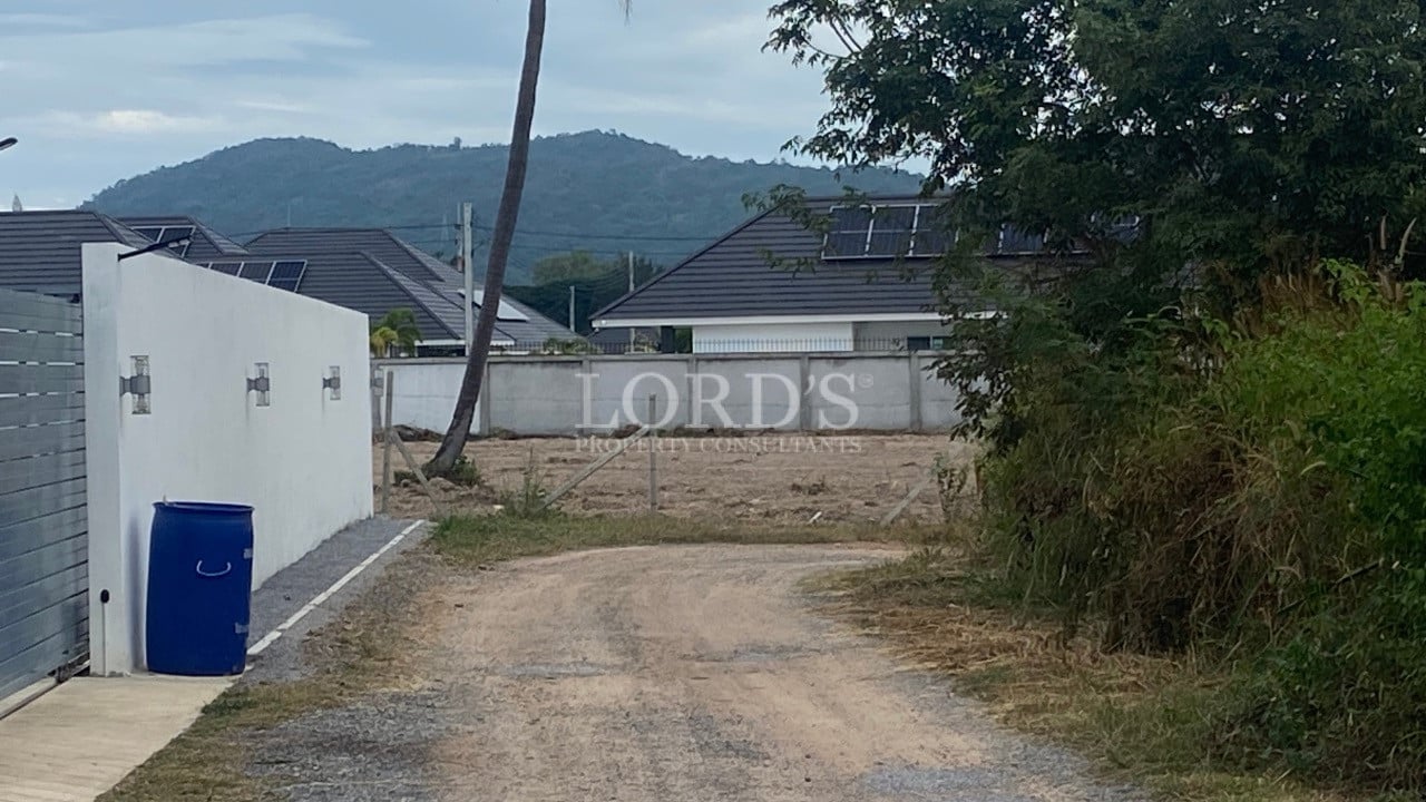 Access road leading to a cleared land plot beside a white boundary wall with mountain views.