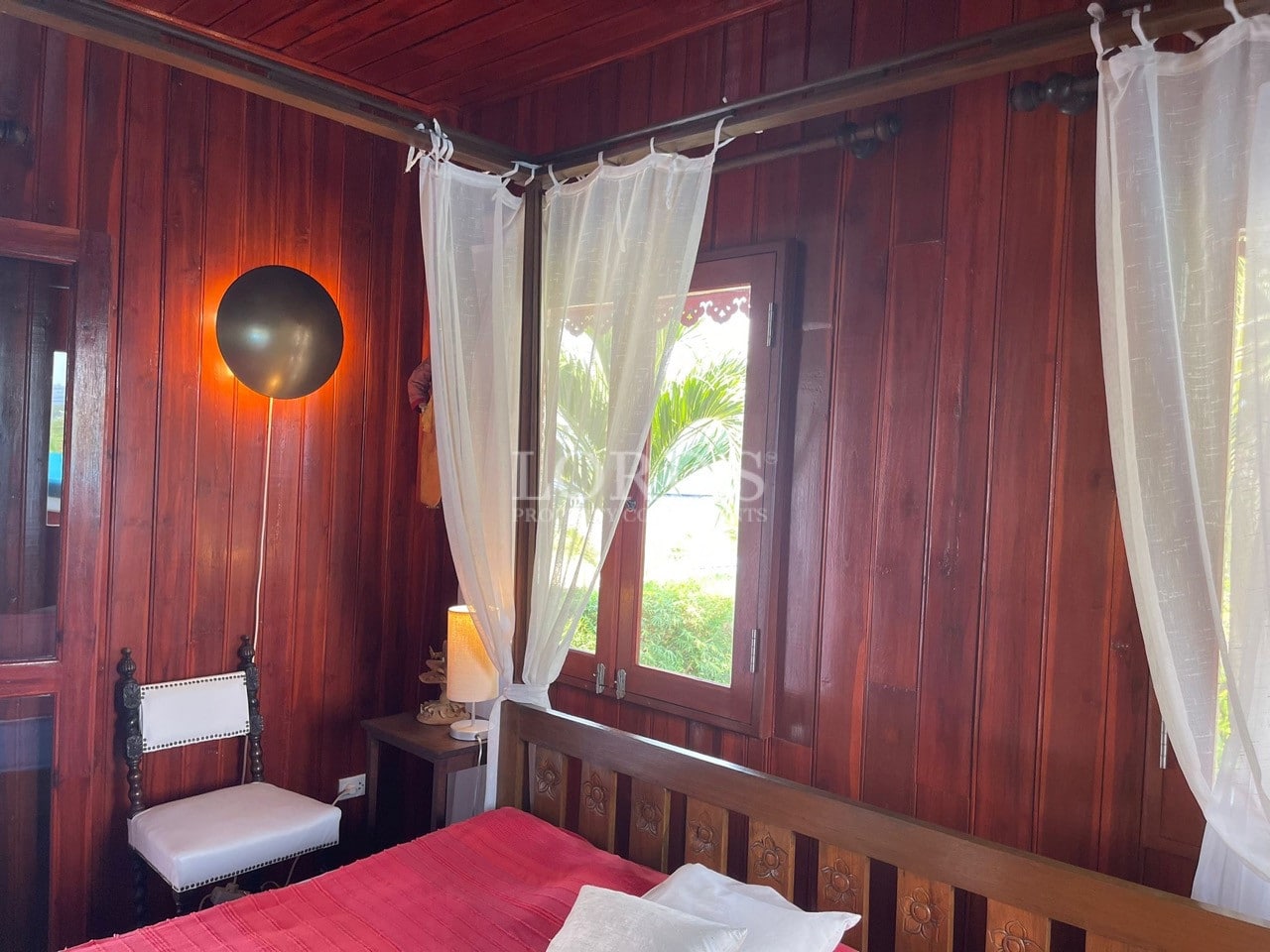 Wooden bedroom interior with canopy bed, sheer curtains, red bedspread, and garden view window.