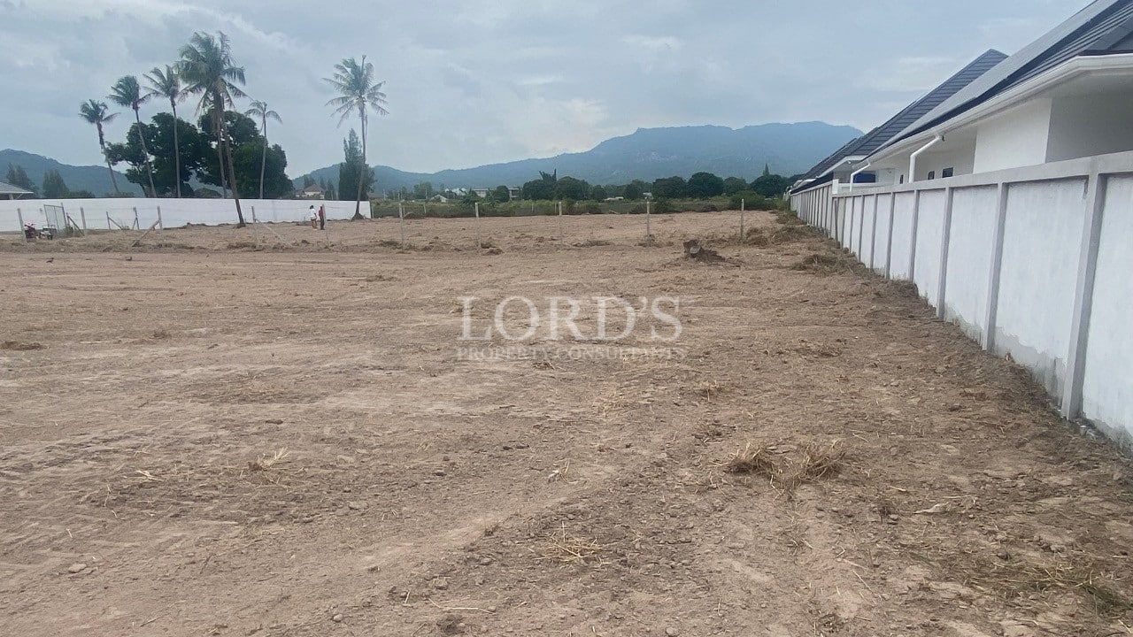 Cleared vacant land plot beside a white boundary wall with mountain views in the background.