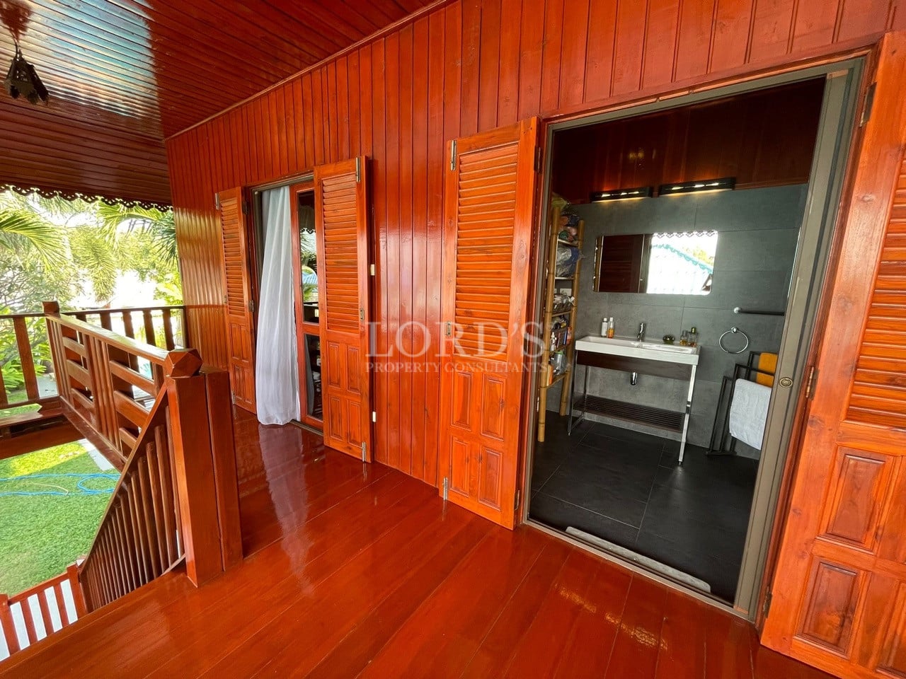 Wooden villa balcony with polished hardwood flooring, shuttered doors, and an open bathroom entrance.