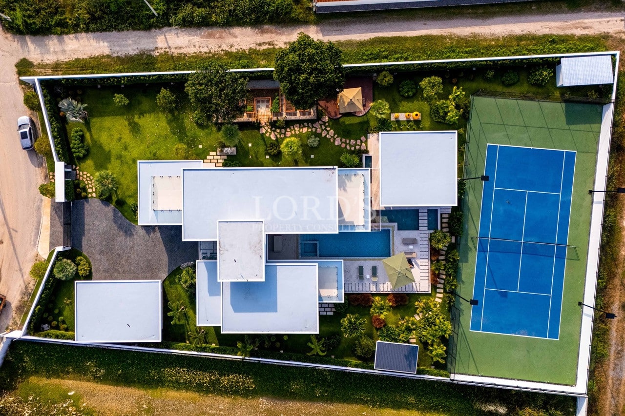 Luftaufnahme einer modernen Luxusvilla mit Swimmingpool, Landschaftsgarten und privatem Tennisplatz aus der Vogelperspektive.