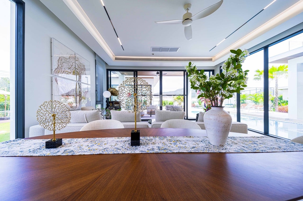 Moderner offener Ess- und Wohnbereich mit Esstisch aus Holz, dekorativem Mittelteil, Deckenventilator und großen Glastüren mit Blick auf den Swimmingpool.