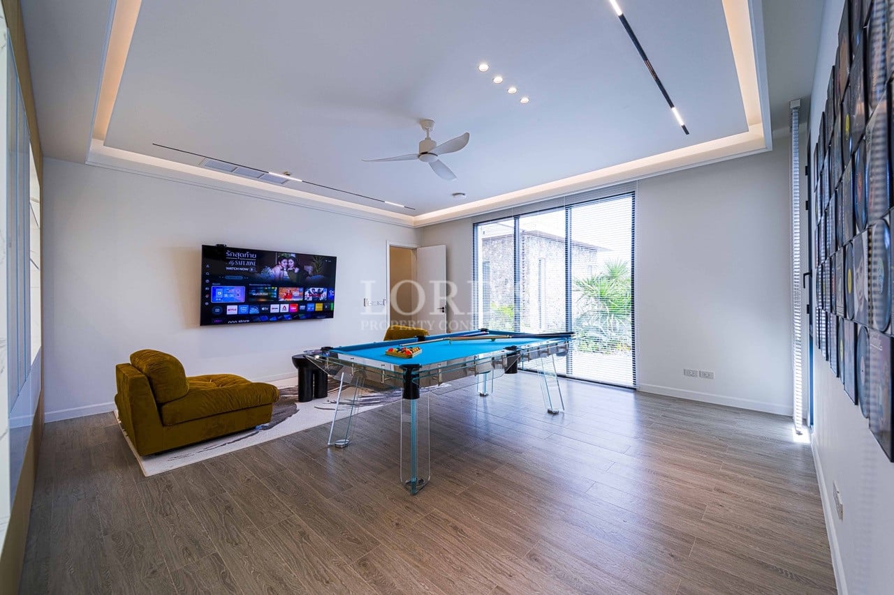 Moderner Unterhaltungsraum mit einem gläsernen Billardtisch, einem an der Wand montierten Smart-TV, einem Sessel und großen Fenstern.