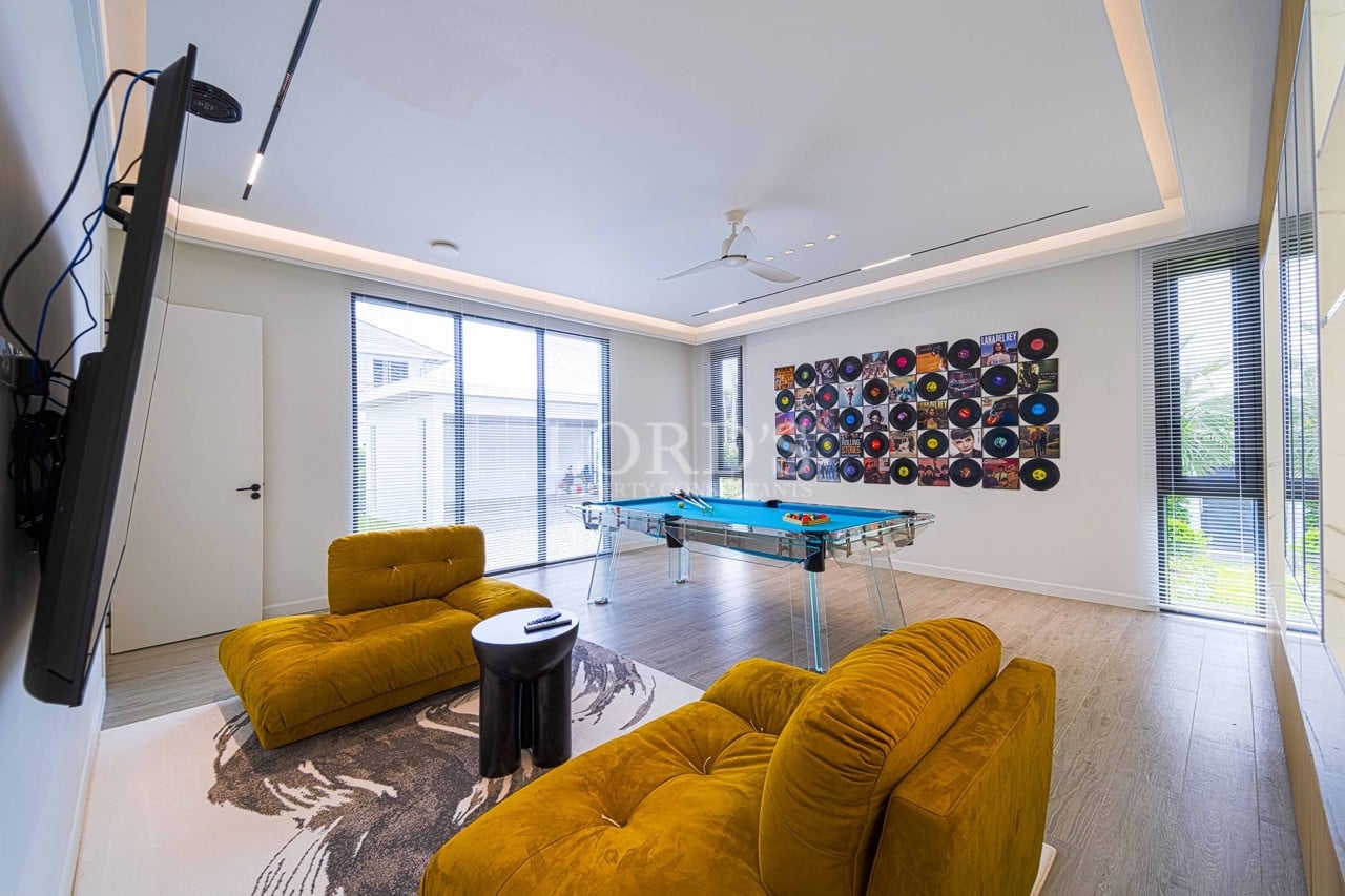 Moderner Unterhaltungsraum mit gläsernem Billardtisch, Vinyl-Schallplatten an der Wand, senfgelben Lounge-Sesseln und raumhohen Fenstern.