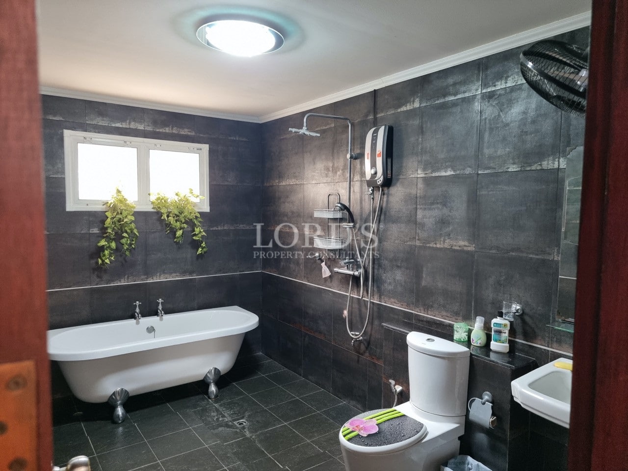Modern bathroom with dark tile walls, freestanding bathtub, rainfall shower, and natural light.