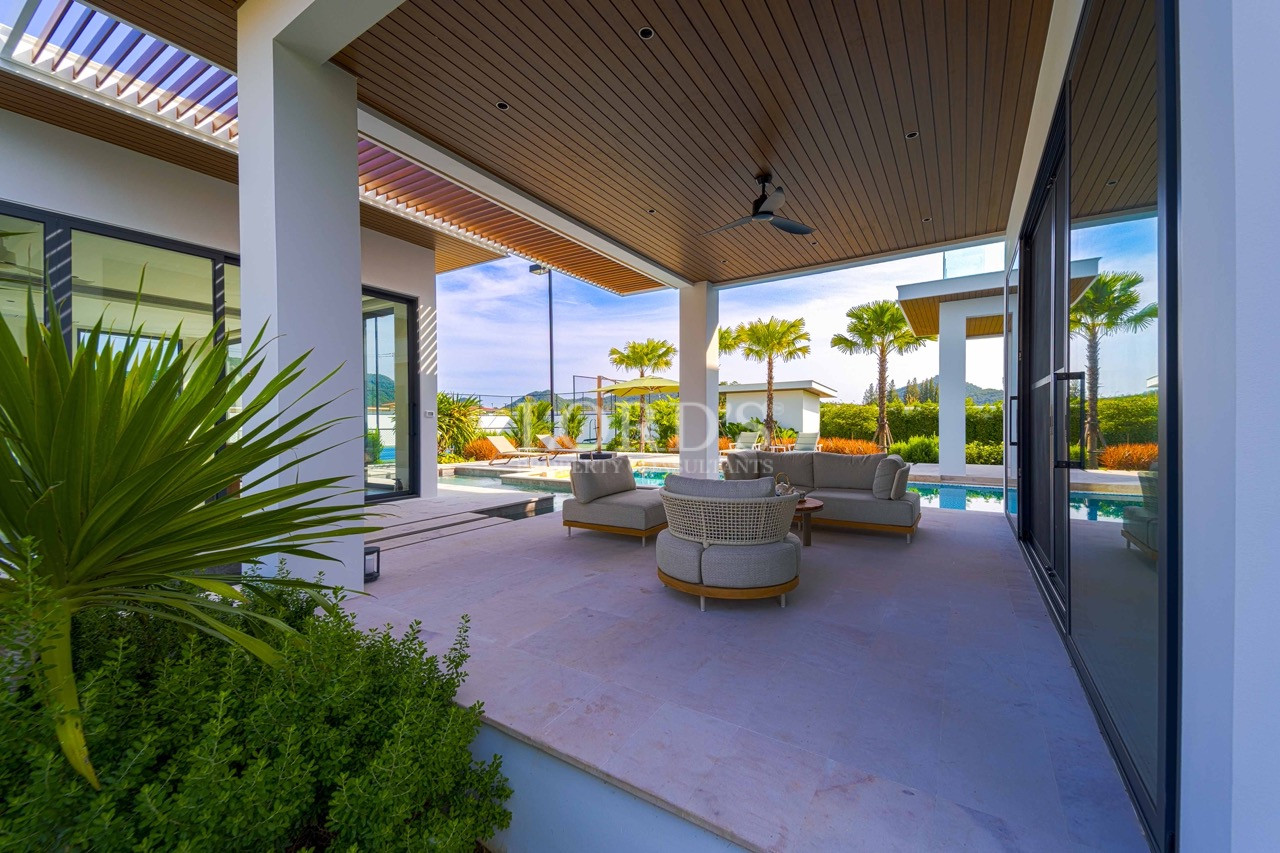 Überdachter Lounge-Bereich im Freien mit modernen Sitzgelegenheiten, Deckenventilator, tropischen Pflanzen und Swimmingpool in einer Luxusvilla.