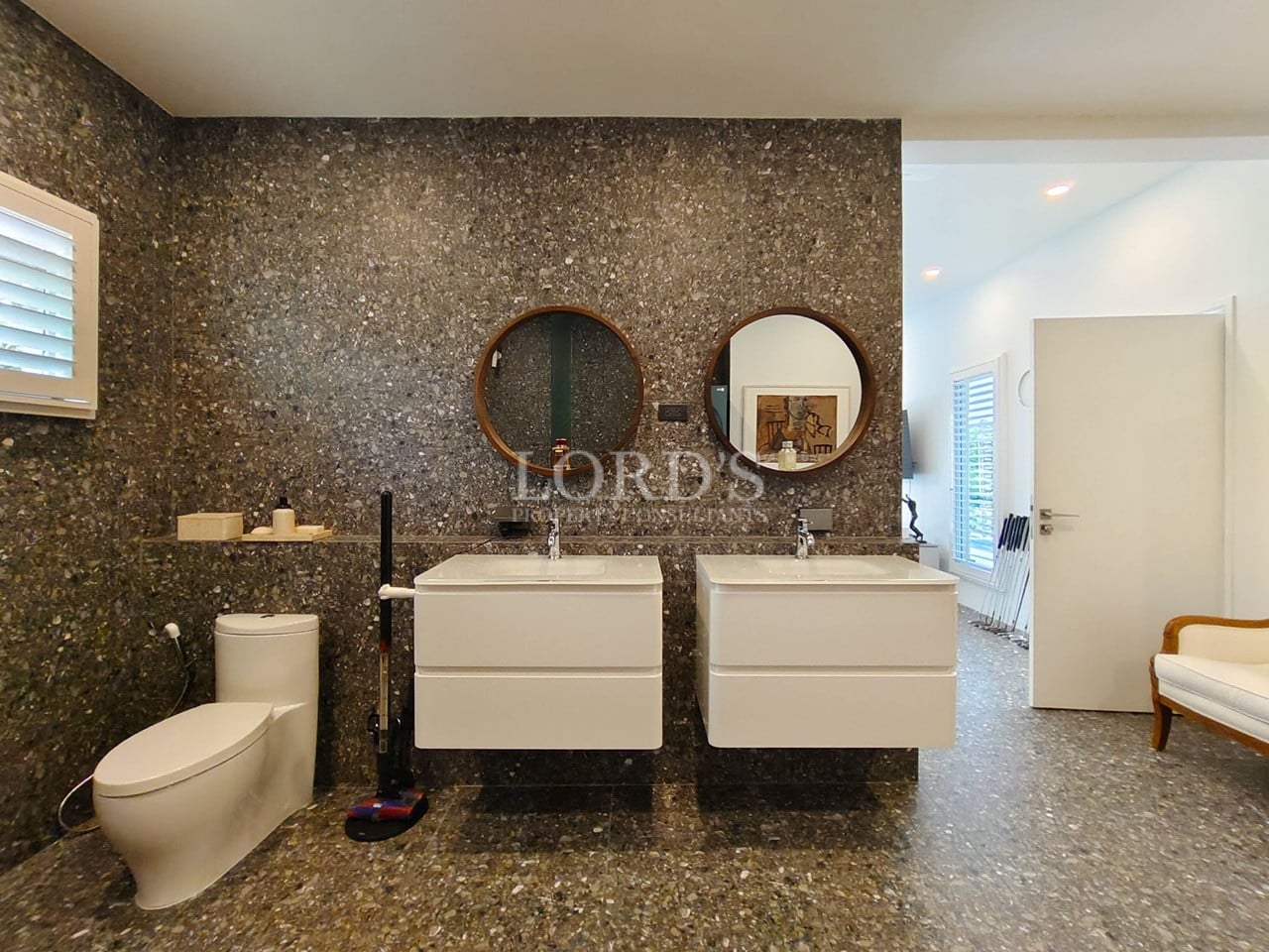 Luxury bathroom with double floating vanity, circular mirrors, stone-textured walls, and modern fixtures.