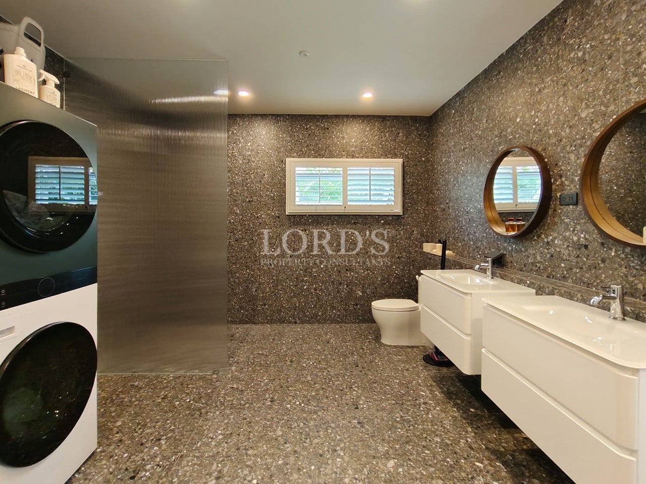 Spacious modern bathroom with double vanity, walk-in shower, stone-textured walls, and integrated washer and dryer.