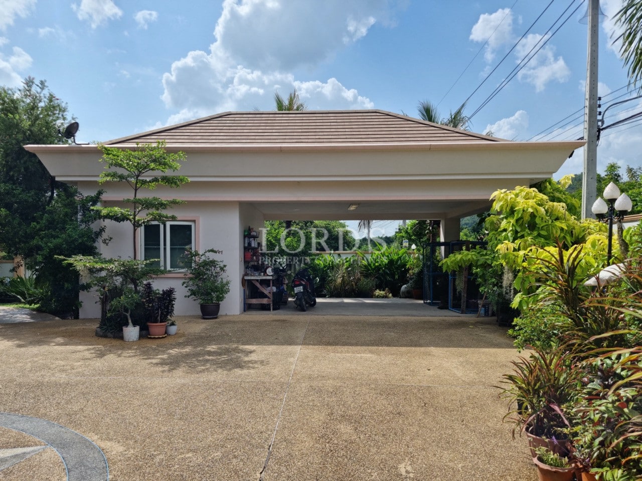 Modern villa with a covered carport and wide concrete driveway surrounded by tropical landscaping.