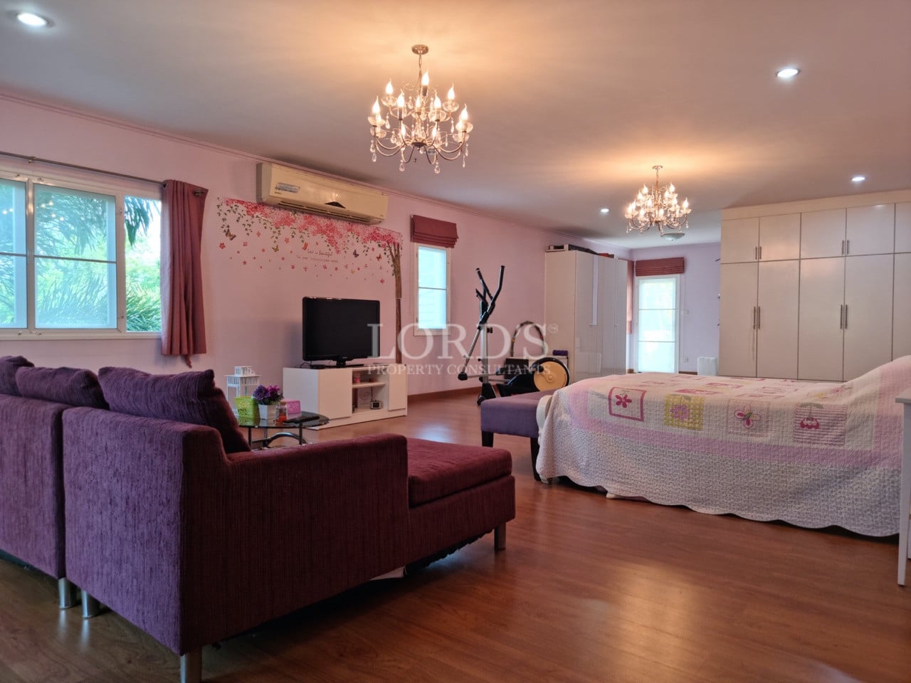 Spacious bedroom with wooden flooring, crystal chandeliers, purple sofa seating, and built-in wardrobe.