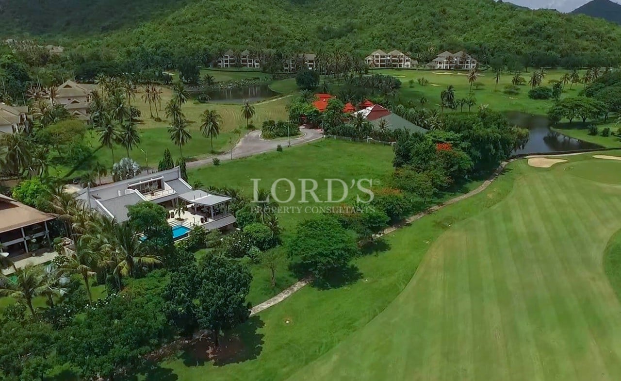 Aerial view of a luxury villa surrounded by a lush green golf course, palm trees, and tropical landscape.