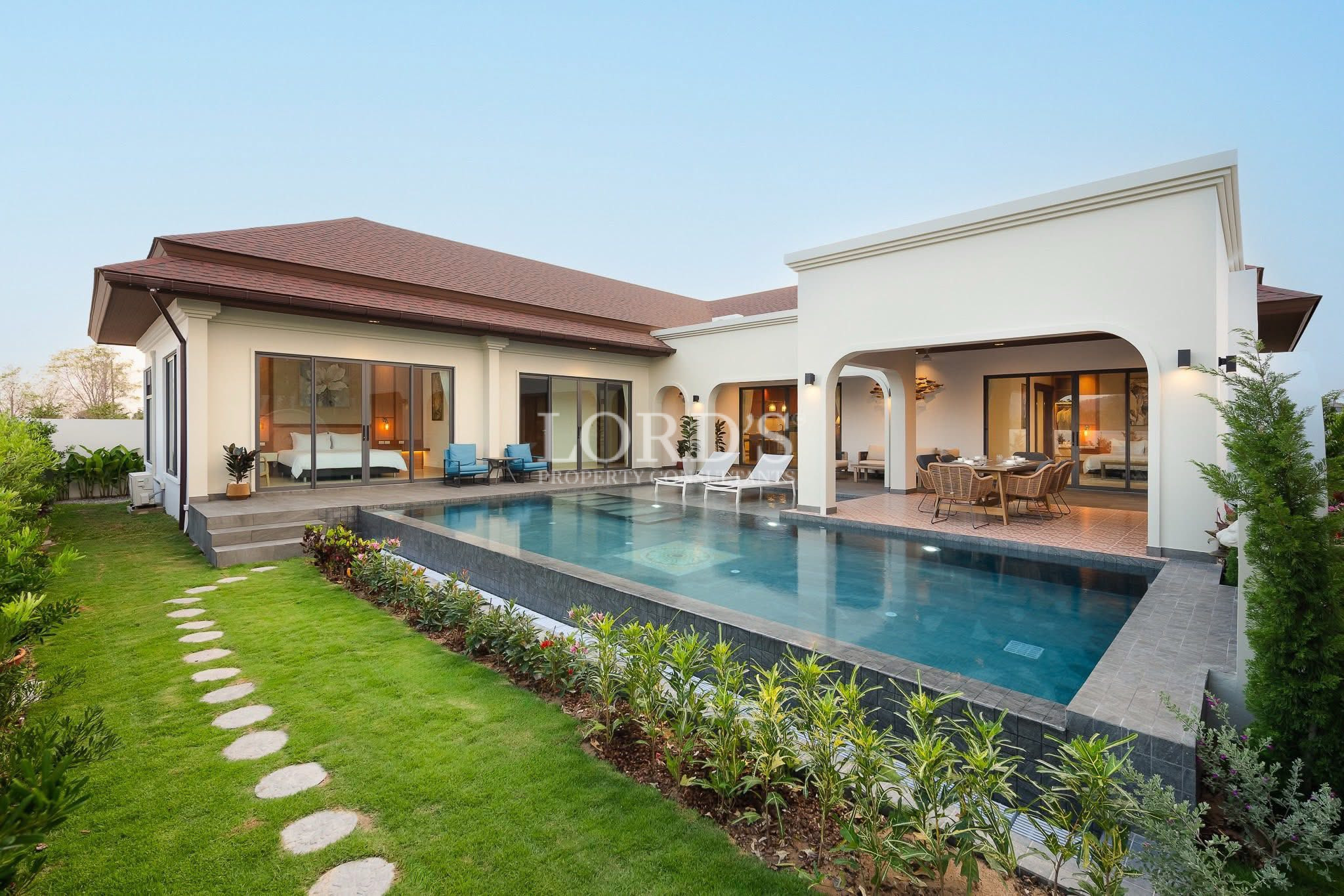 Modern single-story villa with a tiled roof, large glass doors, and a rectangular swimming pool bordered by a manicured lawn and patio seating.