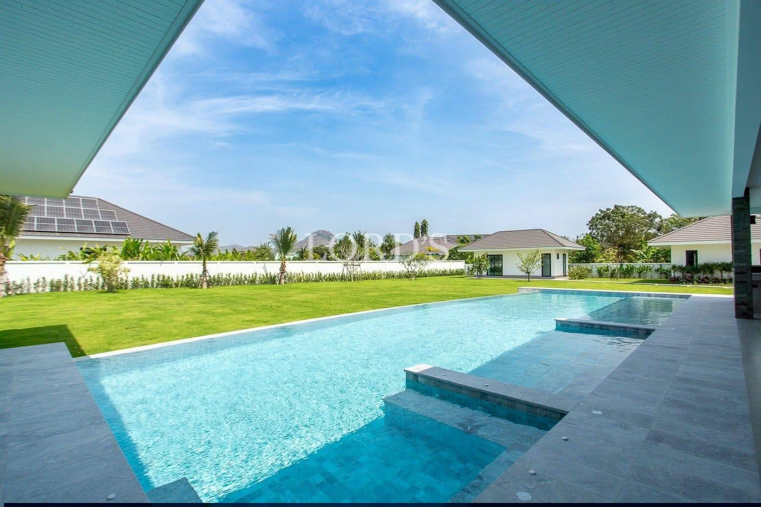 Modern backyard with a large rectangular swimming pool, green lawn, and small white guesthouses under a blue sky.
