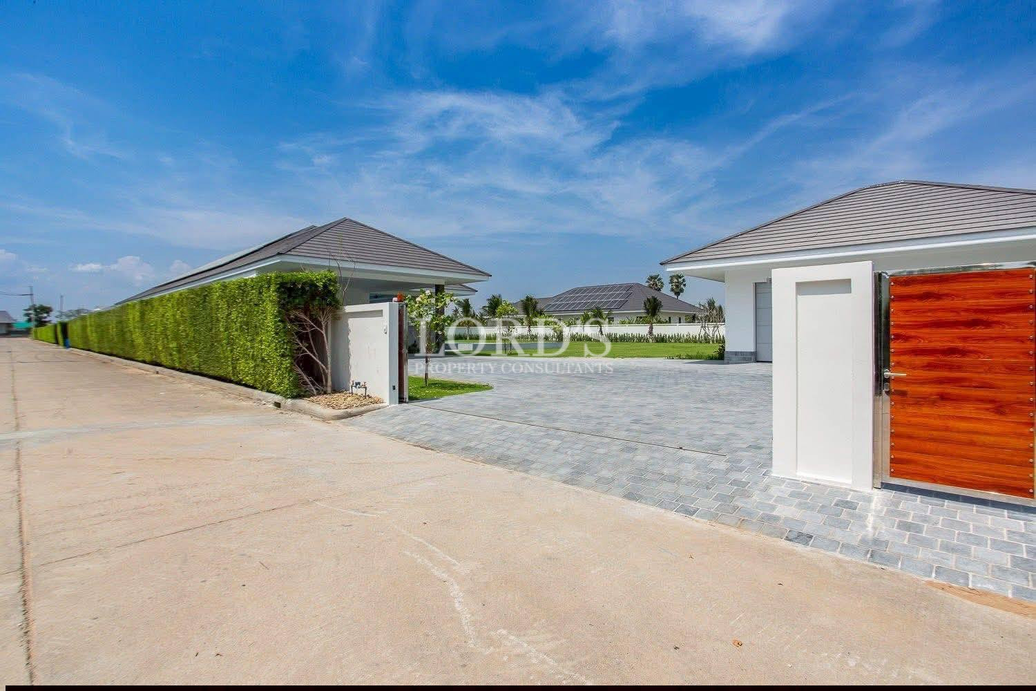 Modern luxury villa entrance with wooden gate, long hedge fence, and paved driveway