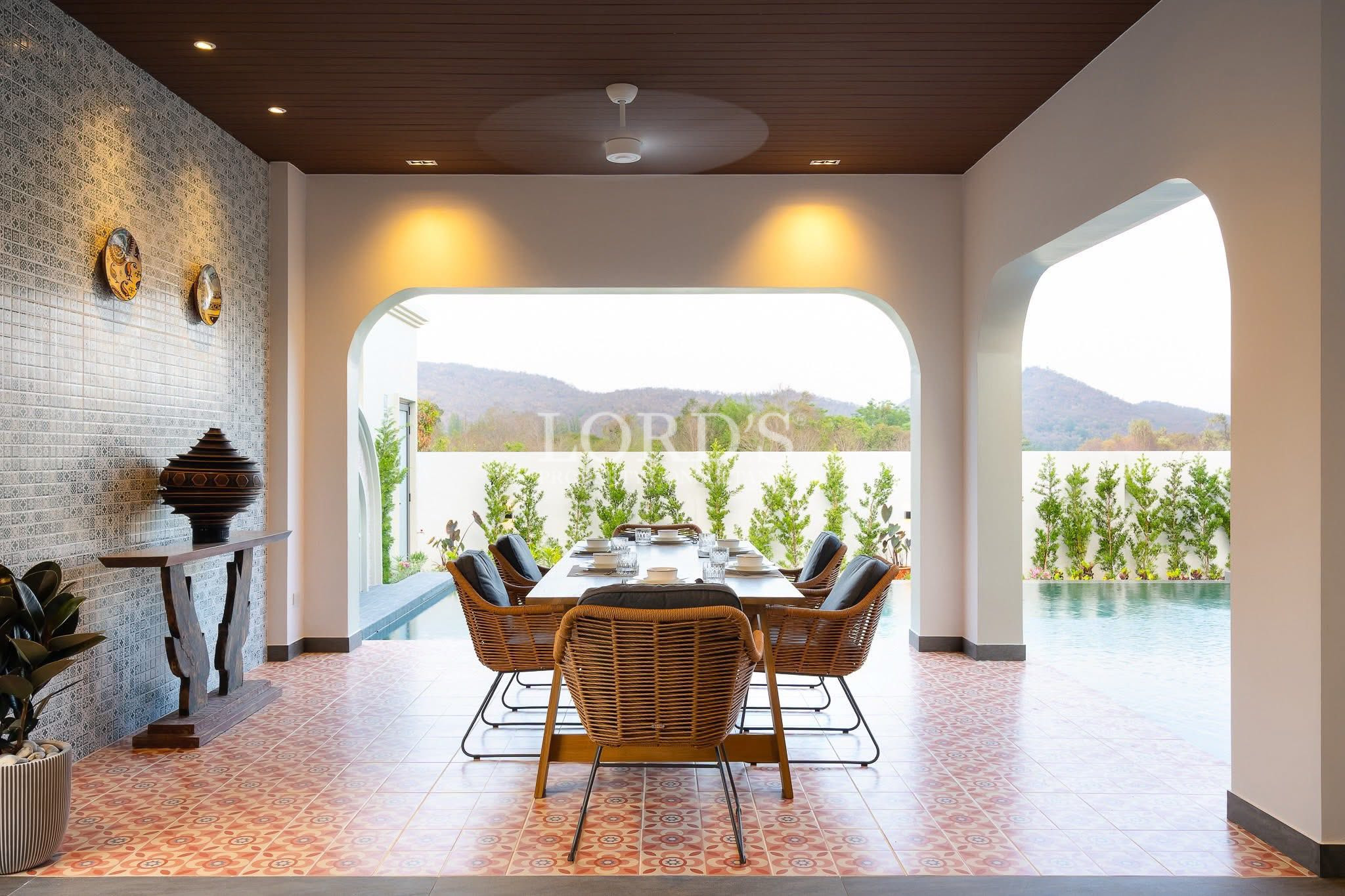 Covered outdoor dining area with wicker chairs overlooking a private pool and garden.