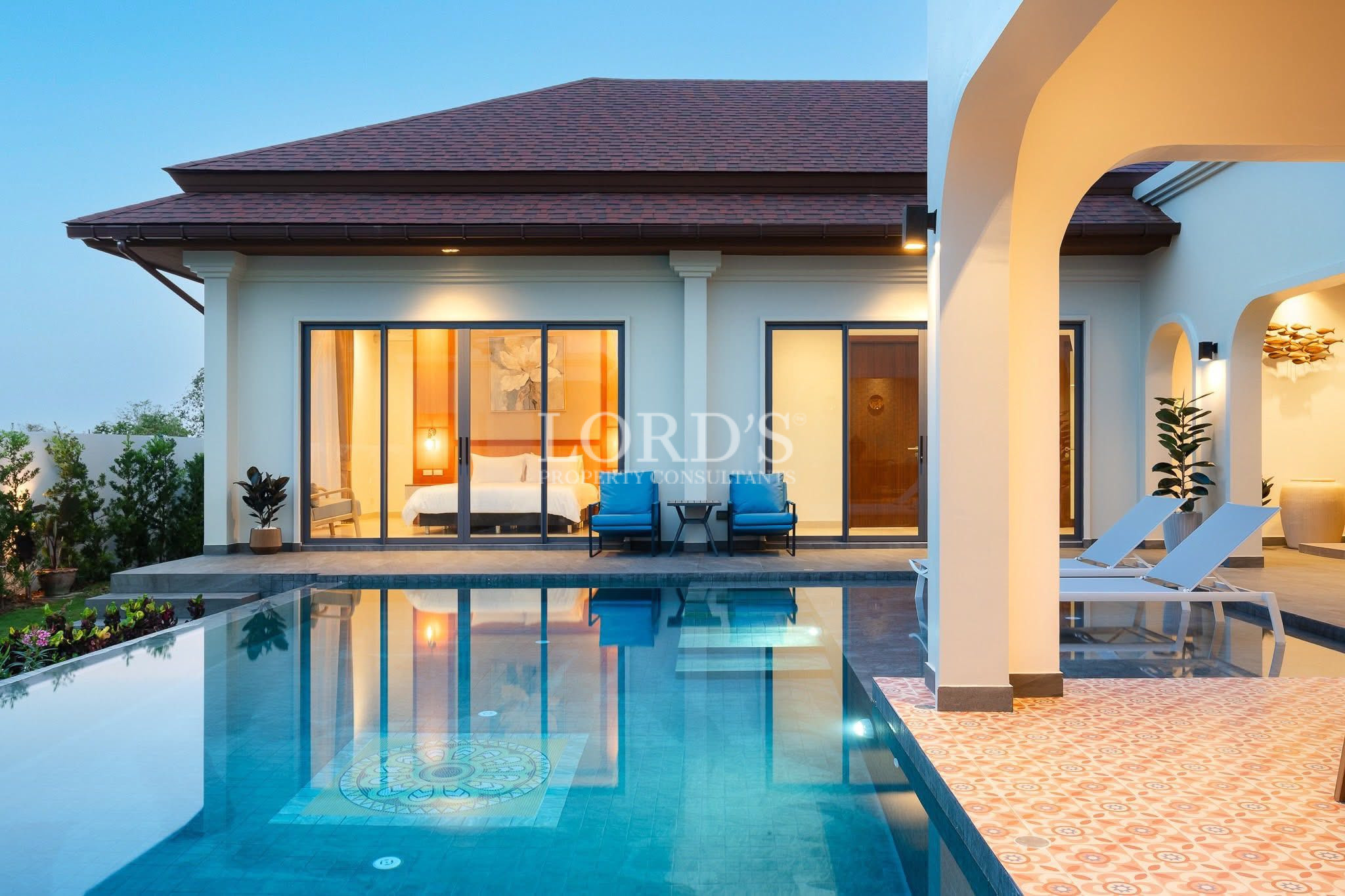 Private villa pool with bedroom view, patio arches, and outdoor lounge chairs at dusk.