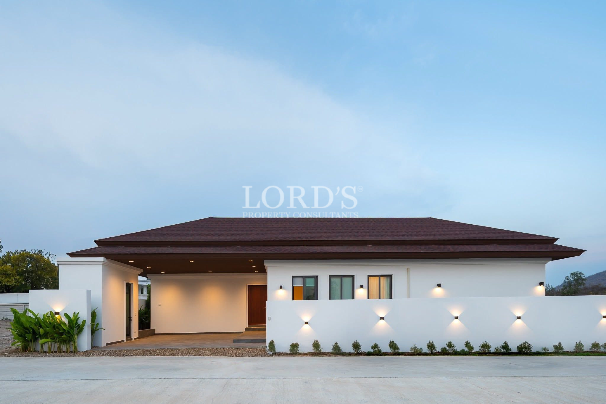 Modern single-story villa exterior with covered carport and illuminated perimeter wall at dusk.