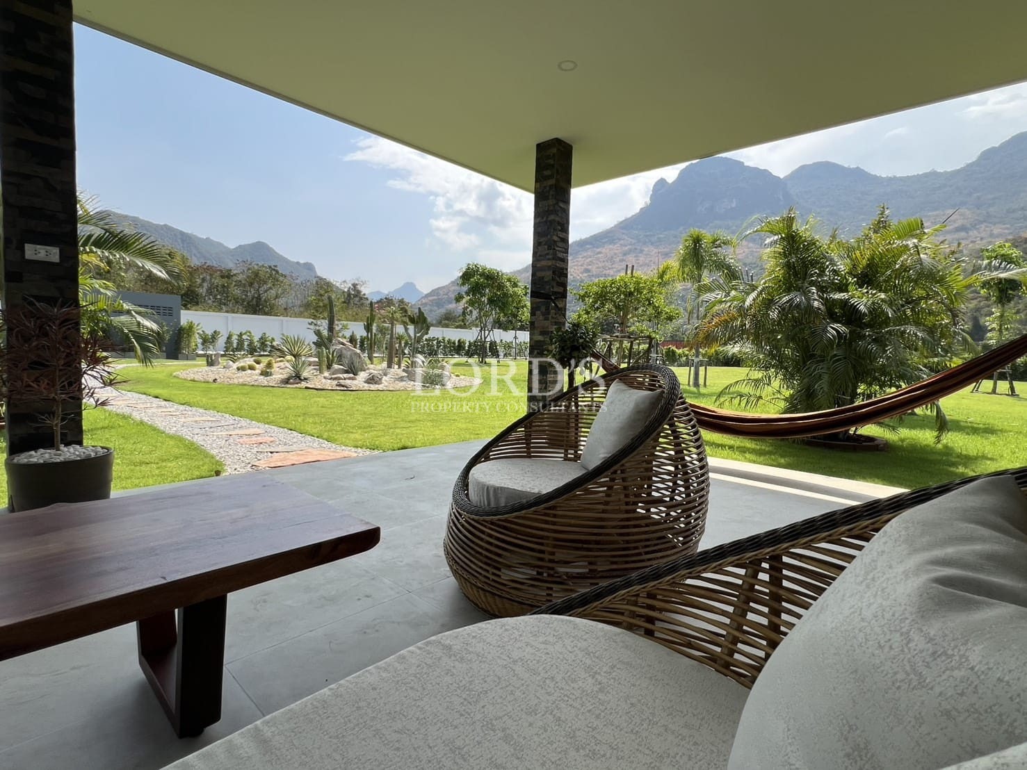 Covered outdoor lounge area with wicker seating, garden landscape, and mountain views.