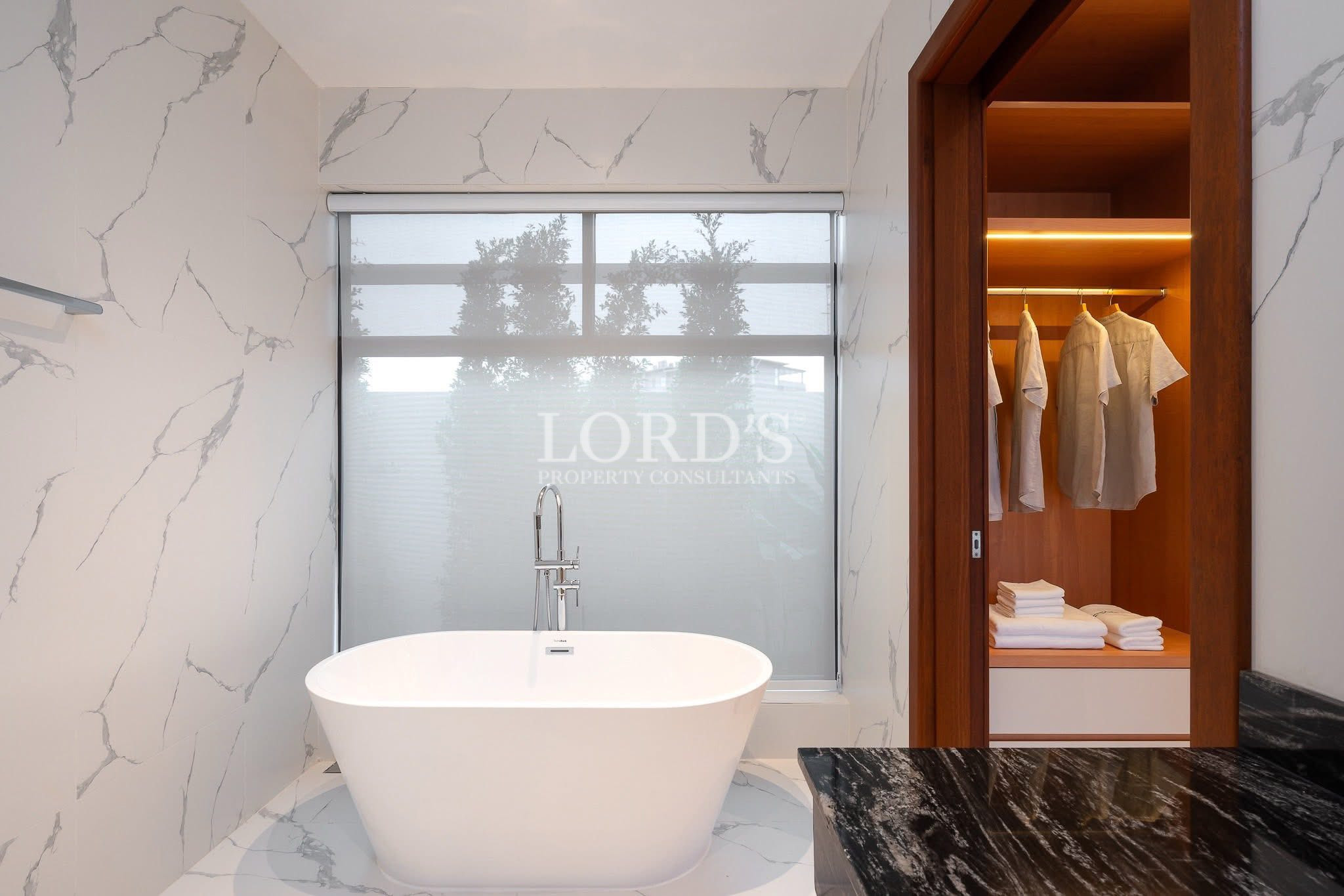 Freestanding bathtub in a modern marble bathroom beside an illuminated walk-in closet.