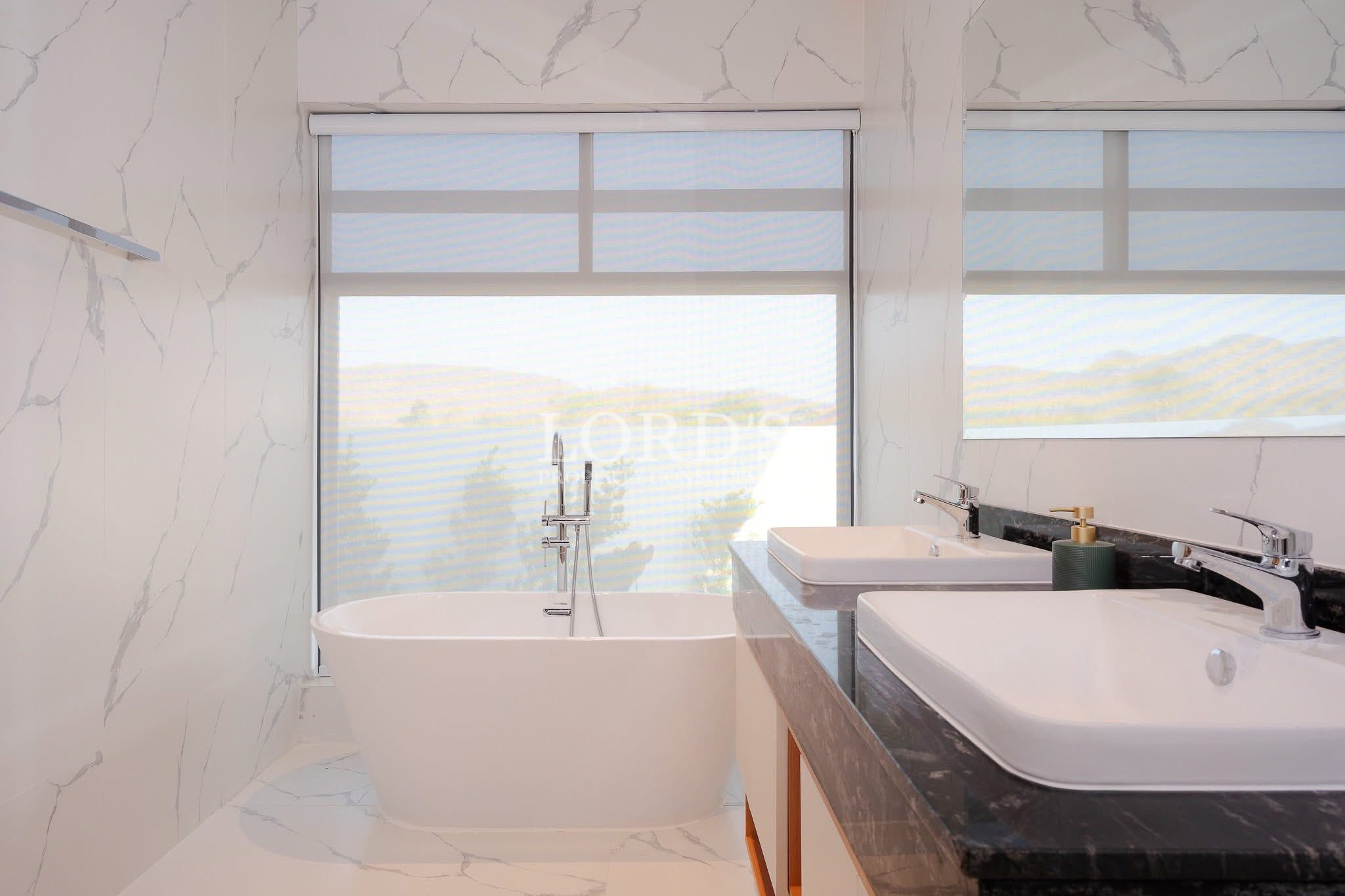 Modern bathroom with a freestanding bathtub in front of a large window and a double-sink vanity with marble-style walls.