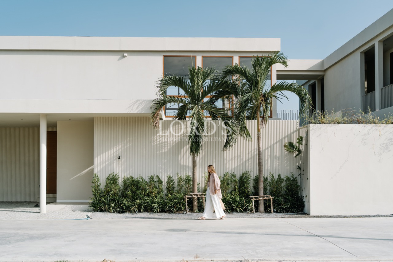Woman walking past a modern white villa with palm trees and minimalist landscaping.