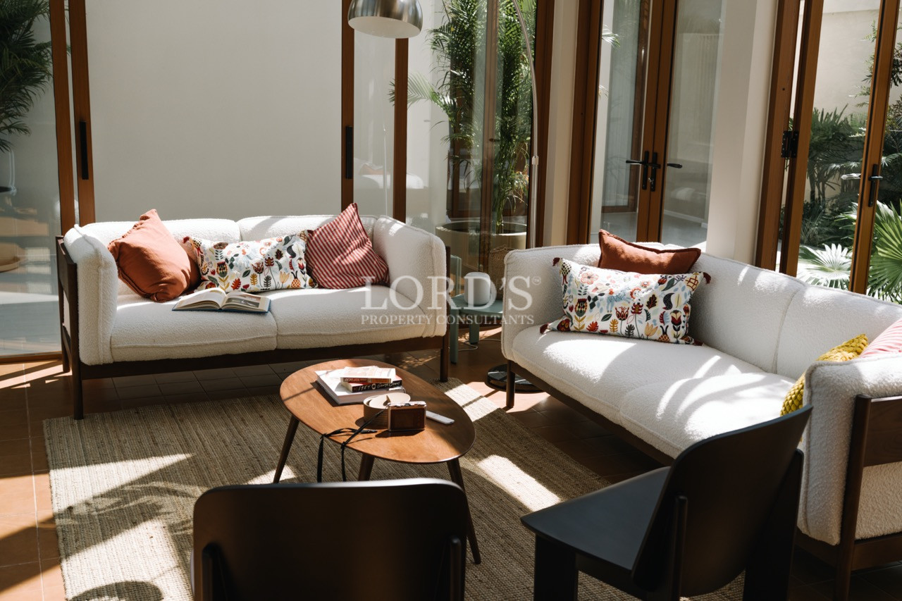 Sunlit living room with two cream sofas, patterned cushions, and a wooden coffee table.