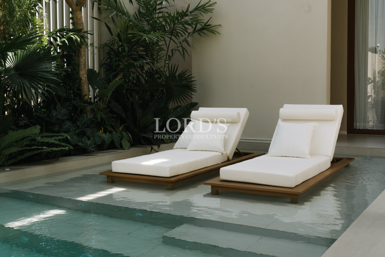 Two white cushioned sun loungers on a shallow ledge beside a private pool surrounded by tropical plants.