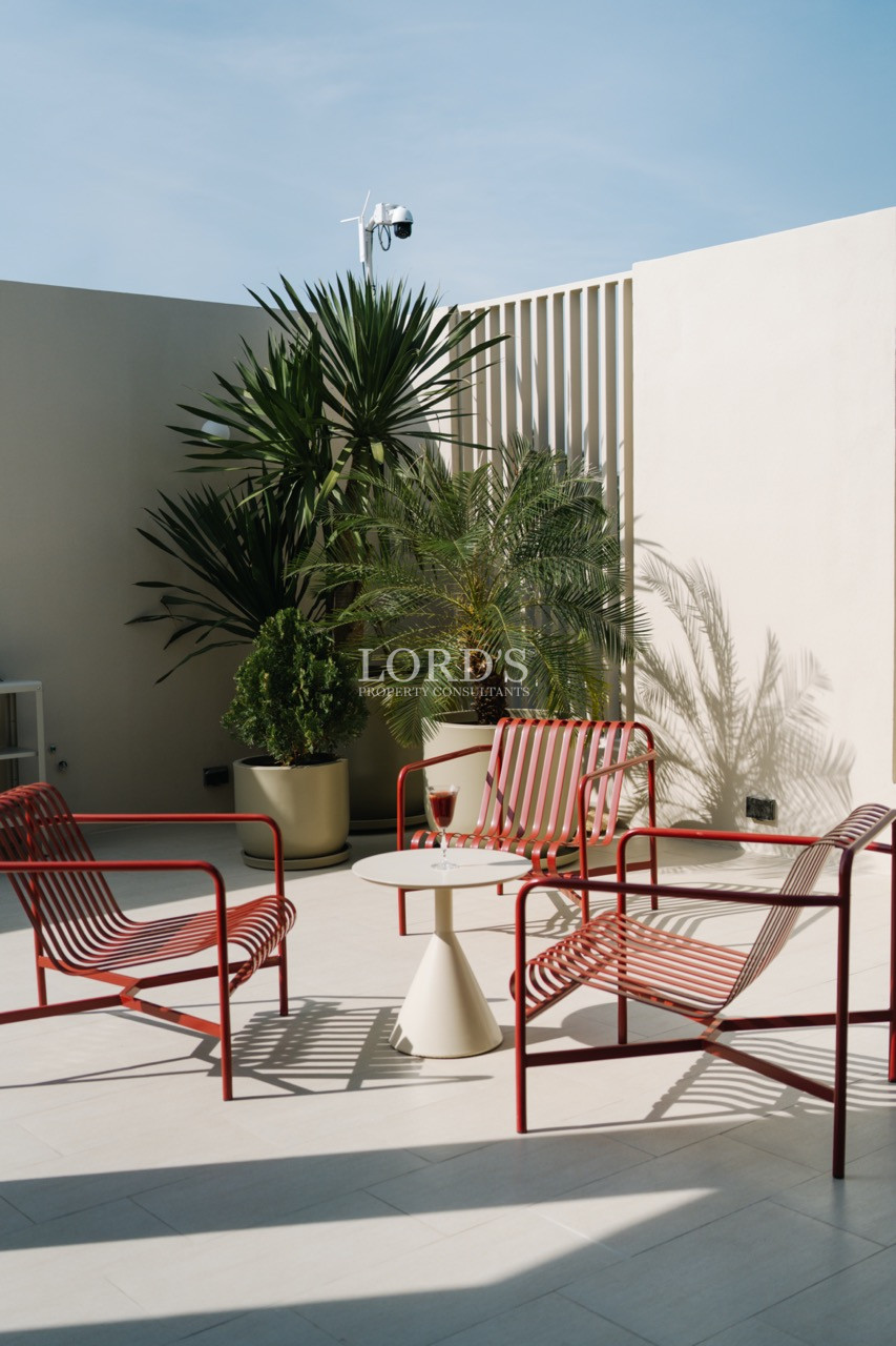 Outdoor terrace with red metal lounge chairs, small round table, and potted plants against a modern white wall.