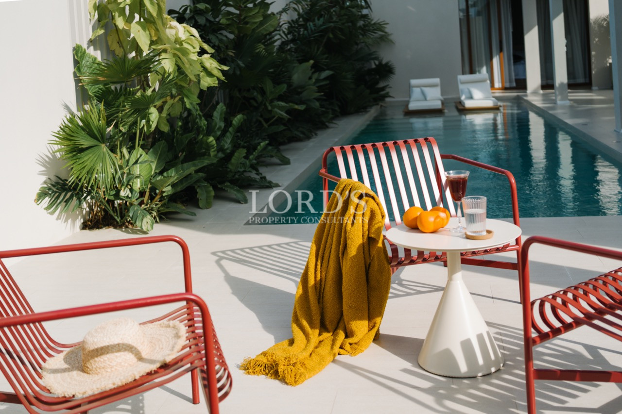 Outdoor poolside seating area with red chairs, small table, drinks, and lush tropical plants.