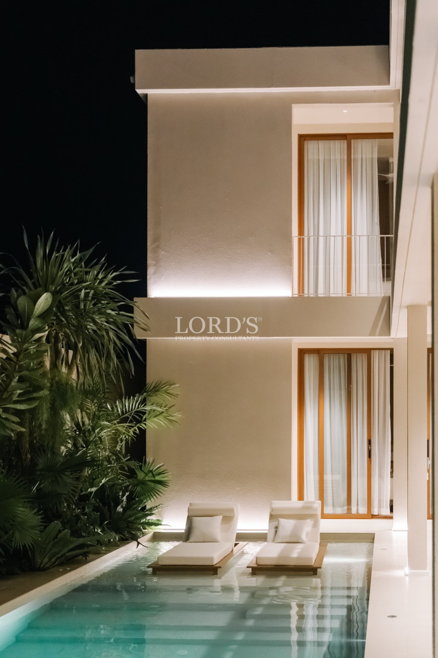 Modern two-story villa at night with illuminated exterior walls, narrow swimming pool, and two white lounge chairs surrounded by tropical plants.