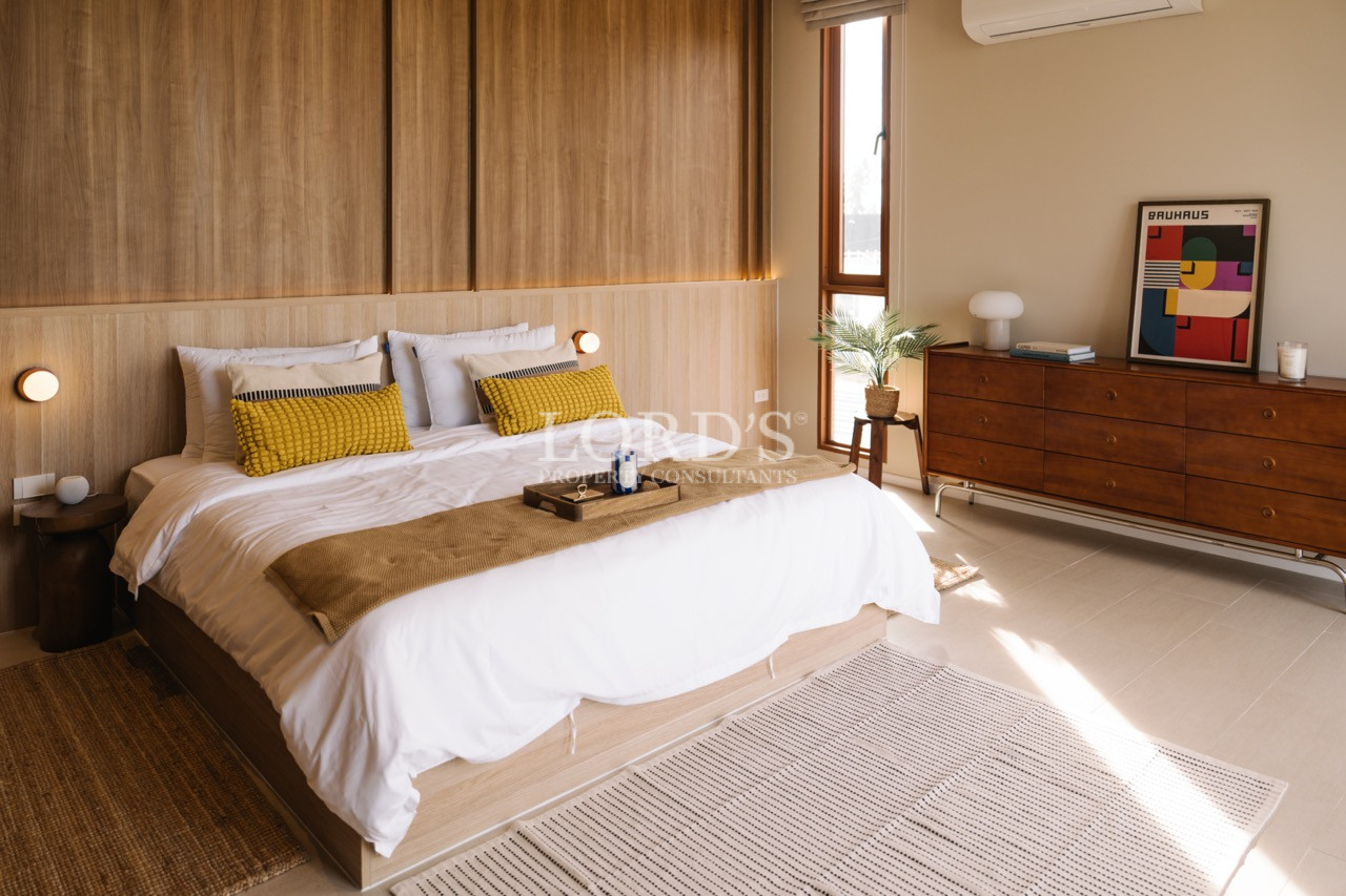 Modern bedroom with a wooden accent wall, double bed with white bedding and mustard cushions, and a mid-century style dresser beside a tall window.
