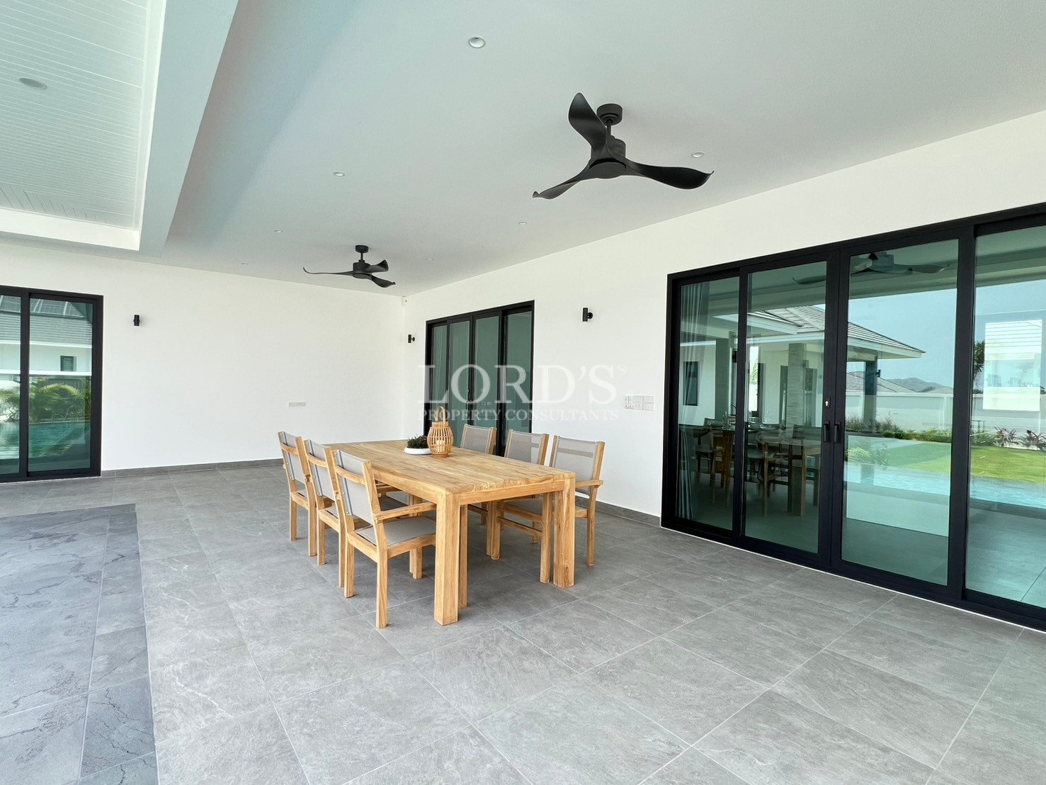 Covered outdoor patio with wooden dining table, ceiling fans, and large sliding glass doors.