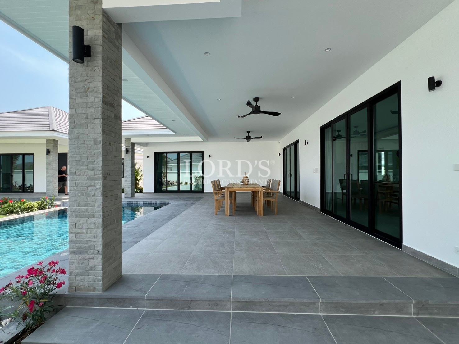 Modern luxury villa patio with wooden dining table, ceiling fans, sliding glass doors, and swimming pool.