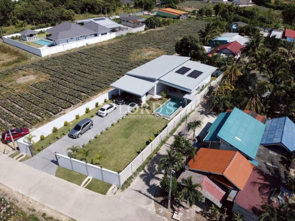 Aerial view of a modern villa with solar panels, swimming pool, driveway, and landscaped lawn surrounded by farmland and neighboring homes.