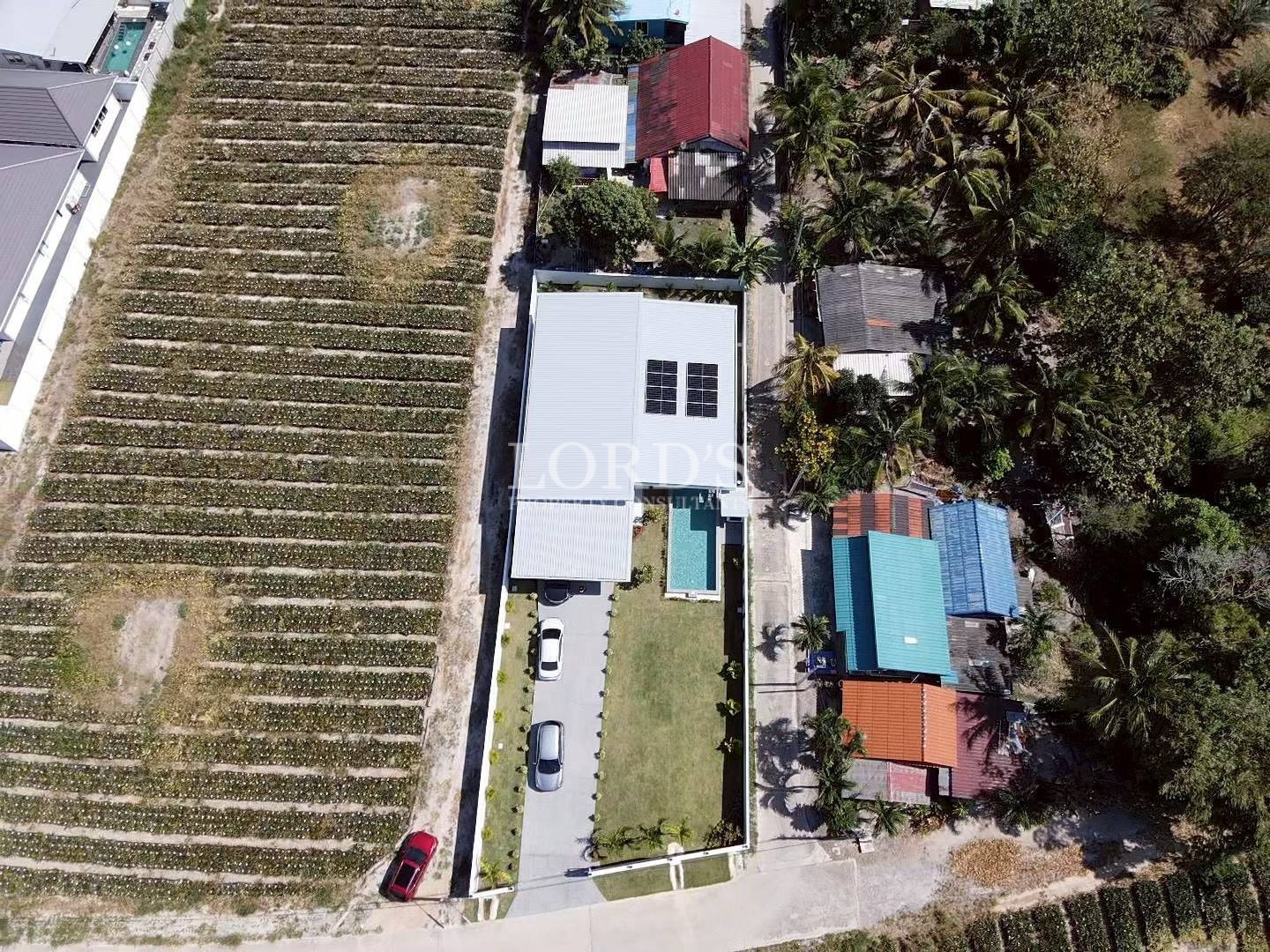 Aerial view of a modern home with solar panels, driveway, lawn, and swimming pool surrounded by neighboring houses and farmland.